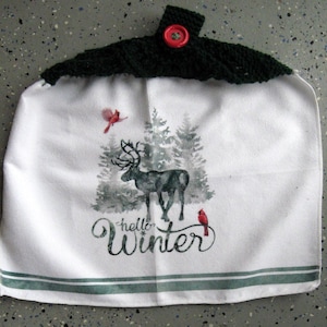 May include: White kitchen towel with a winter scene featuring a deer, pine trees, and red birds. The towel has a green crocheted top with a red button and the words "hello Winter". Green stripes are at the bottom.
