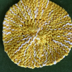 May include: A round, hand-knitted washcloth in a spiral pattern. The washcloth is primarily yellow with white stripes, creating a textured appearance. The item is displayed on a green surface.