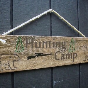 May include: A rustic wooden sign with the text "Hunting Camp" in brown lettering. The sign is decorated with green pine trees, a rifle, a bear, and a deer. The sign is hanging from a white rope.