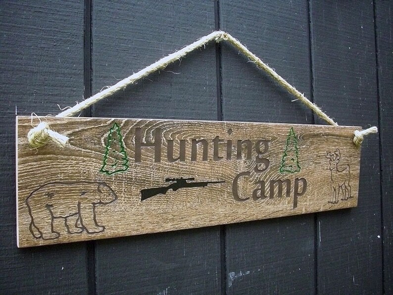 May include: A rustic wooden sign with the text "Hunting Camp" in brown lettering. The sign is decorated with green pine trees, a rifle, a bear, and a deer. The sign is hanging from a white rope.