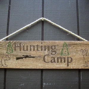 May include: A wooden sign with the text "Hunting Camp" in brown lettering. The sign is decorated with a silhouette of a bear, a rifle, a deer, and two pine trees.