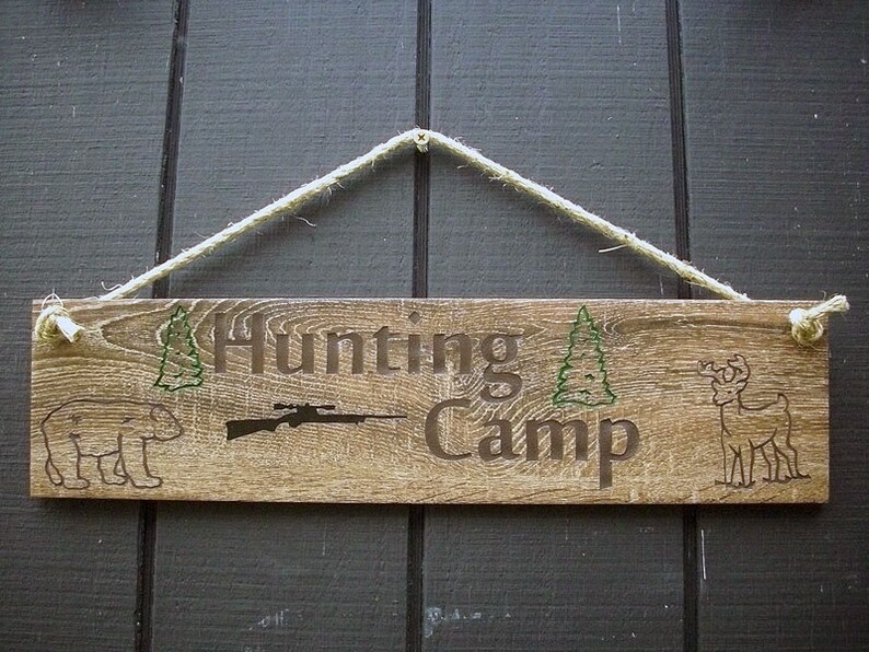 May include: A wooden sign with the text "Hunting Camp" in brown lettering. The sign is decorated with a silhouette of a bear, a rifle, a deer, and two pine trees.