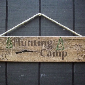 May include: A rustic wooden sign with the text "Hunting Camp" in brown lettering. The sign is decorated with a silhouette of a bear, a rifle, a pine tree, and a deer. The sign is hanging from a rope.