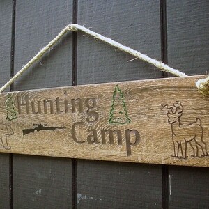 May include: A rustic wooden sign with the text "Hunting Camp" in brown lettering. The sign also features a silhouette of a rifle, a deer, and two pine trees. The sign is hanging from a rope.