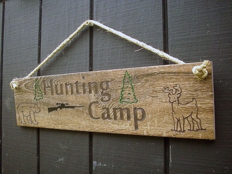 May include: A rustic wooden sign with the text "Hunting Camp" in brown lettering. The sign also features a silhouette of a rifle, a deer, and two pine trees. The sign is hanging from a rope.