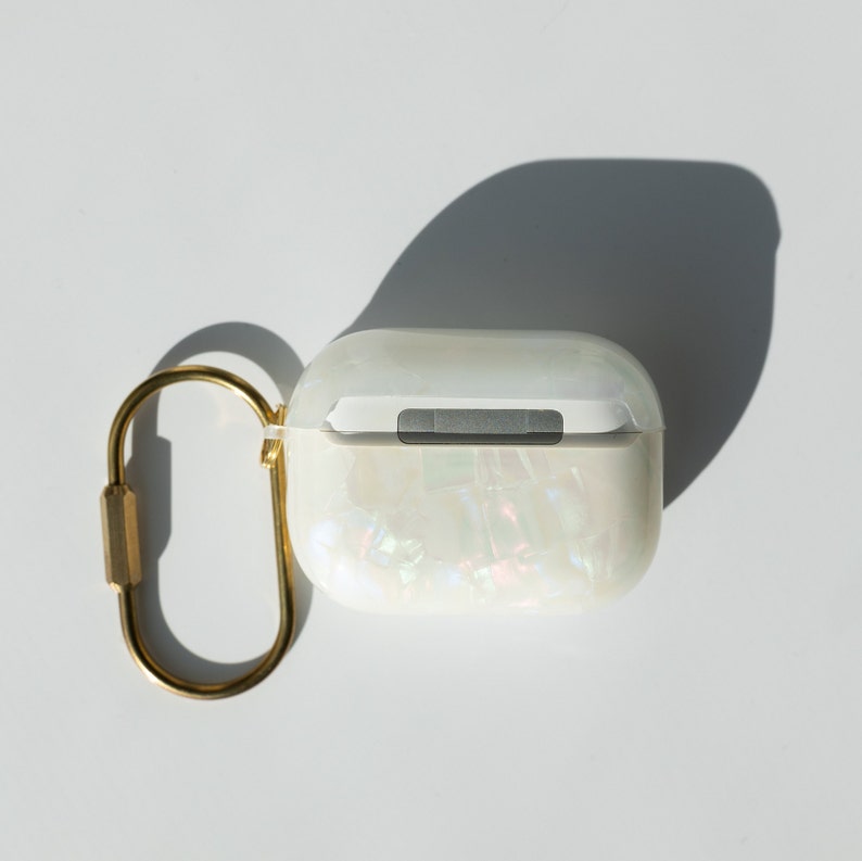 Może przedstawiać: Białe, perłowe etui na słuchawki Airpods z złotym metalowym k&oacute;łkiem do kluczy.
