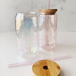 May include: Two iridescent glass can-shaped tumblers with bamboo lids and pink reusable straws. One tumbler has a straw inserted through the lid.