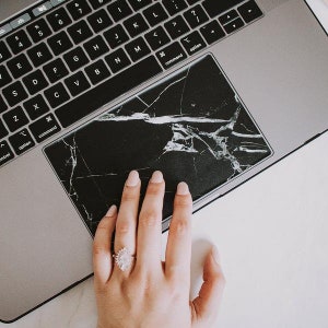 May include: A laptop computer with a black marble-patterned trackpad skin. The skin has a cracked effect, resembling broken glass.