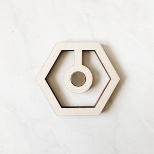 May include: A wooden hexagonal frame with a circular cutout in the centre. The frame is light brown and has a slightly distressed finish. A small, round, silver magnet is visible in the centre of the cutout.