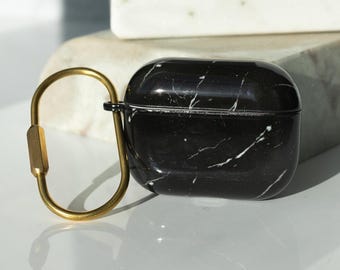May include: Black marble-patterned AirPod case with a gold-colored carabiner clip. The case has a glossy finish and white veining. The carabiner is attached to the case with a small black hinge. The background is a white surface.