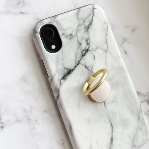 May include: A white marble phone case with a gold ring holder. The ring holder has a white center.