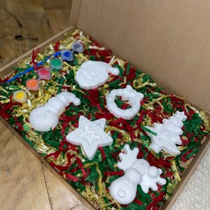 May include: A set of six white ceramic Christmas decorations in a box with red, green and gold shredded paper. The decorations are shaped like a snowman, a star, a reindeer, a Christmas tree, a Santa Claus and a candy cane.