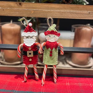 May include: Two festive Christmas tree ornaments: a Santa Claus and an elf. The Santa is red with a white beard and hat, and a black belt. The elf is green with a red collar and hat. Both have candy cane legs.