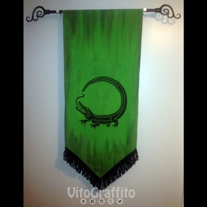 Puede incluir: Un estandarte verde con un diseño de cocodrilo negro. El cocodrilo está enroscado en un círculo y tiene su cola en la boca. El estandarte tiene un fleco negro alrededor de los bordes.