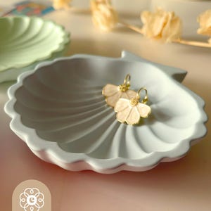 May include: A light blue seashell-shaped trinket dish with a pair of flower earrings. The earrings have a gold-colored hoop and a cream-colored flower design. The dish has a scalloped edge and the text "Chéri Design Lab".