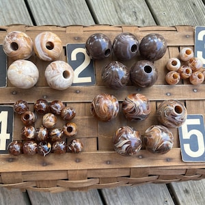 May include: Assortment of brown and cream-colored beads with various patterns, including swirls and marbling, displayed on a woven basket. The beads are of different sizes and shapes, with some having holes. Numbered cards are placed near the beads.