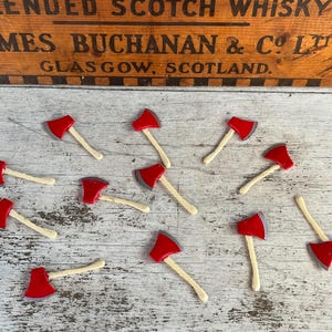 May include: A collection of miniature axes with red axe heads and light-colored wooden handles are scattered on a wooden surface. The axes are arranged in a random pattern. In the background, a wooden sign reads "BLENDED SCOTCH WHISKY JAMES BUCHANAN & Co Ltd GLASGOW, SCOTLAND."