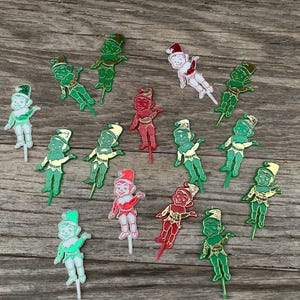 May include: A collection of vintage Christmas elf ornaments in various colors, including green, red, and white. The elf figures are depicted in festive attire, with hats and suits, and are mounted on small sticks.