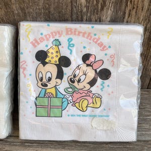 May include: White paper napkins featuring Mickey and Minnie Mouse with the text "Happy Birthday." Mickey wears a party hat and holds a gift. Minnie wears a pink bow and a pacifier. The napkins are wrapped in clear plastic.