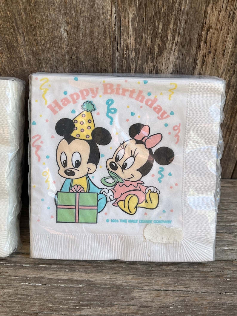 May include: White paper napkins featuring Mickey and Minnie Mouse with the text "Happy Birthday." Mickey wears a party hat and holds a gift. Minnie wears a pink bow and a pacifier. The napkins are wrapped in clear plastic.