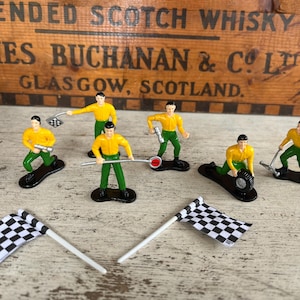 May include: A collection of vintage toy race car figurines. The figurines are dressed in yellow shirts and green pants. Two checkered flags are also present. The background features a wooden box with text that reads "BLENDED SCOTCH WHISKY JAMES BUCHANAN & CO. LTD GLASGOW, SCOTLAND."