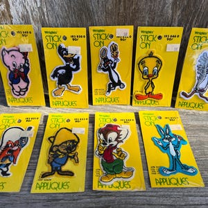 May include: A collection of vintage Wrights Stick-On Appliques featuring cartoon characters like Porky Pig, Daffy Duck, Tweety Bird, and Bugs Bunny. Each applique is on a yellow backing card.