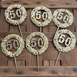 May include: Six gold-colored "50" cake toppers. Each topper features the number "50" in a gold wreath design with a long gold stem. The toppers are arranged on a brown woven basket.