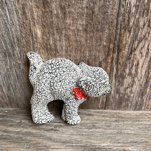 May include: A ceramic cat figurine with a textured black and white speckled pattern. The cat has a small red bow and green eyes. The figurine is standing on a wooden surface.