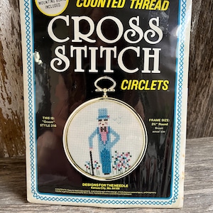 May include: A vintage cross stitch kit featuring a design of a man in a blue suit and top hat, labeled "Groom." The kit includes a gold-tone frame, foam-covered mounting board, and instructions. The frame size is 3.5 inches round.
