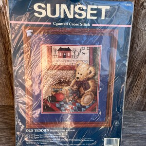 May include: A Sunset counted cross-stitch kit titled "Old Teddies." The kit features a framed design of a teddy bear, a house, and other elements. The packaging includes the Sunset logo and the designer's name, Anna Krajewski.