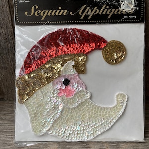 May include: A sequin Santa head applique in a plastic package. The Santa head has a red sequin hat, gold sequin trim, and a white sequin beard. The package has the text "Sequin Appliques".
