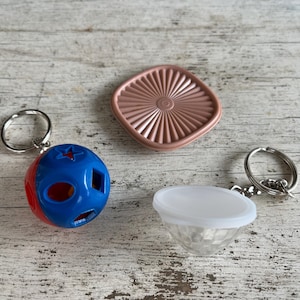 May include: A collection of small, colorful keychains. One is a blue and red ball with cut-out shapes, another is a clear, faceted diamond shape with a white top, and the third is a pink, square-shaped lid.