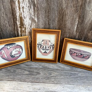 May include: Three framed prints featuring illustrations of pottery. The left print shows a rounded vessel with red and black geometric patterns. The center print displays a vase with black and white floral motifs. The right print depicts a bowl with red and black zig-zag designs. Each print is housed in a rectangular wooden frame with a multi-layered, textured border.