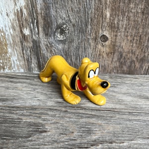 May include: A yellow ceramic figurine of Pluto, a Disney cartoon dog, in a crawling pose. The figurine has a black collar with a red tag, black nose, and white eyes. The background is a weathered wooden surface.