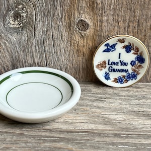 May include: Two small, white ceramic dishes. One is oval with a green rim and a green inner circle. The other is round with a gold rim and the words "I Love You Grandma" surrounded by blue flowers and a blue bird.