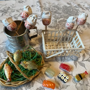 May include: A whimsical still life featuring miniature food items. Includes skewers with chef faces in a metal bucket, a white shopping basket, a woven basket with fish, and various sushi pieces. The scene is set on a floral patterned surface.