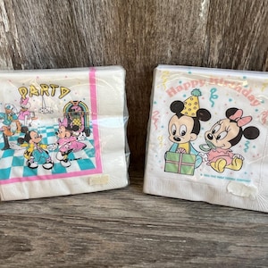May include: Two packs of white paper napkins. One pack features Mickey and Minnie Mouse at a party, with the word "PARTY" at the top. The other pack has Mickey and Minnie with the words "Happy Birthday".