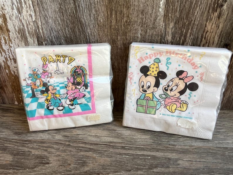 May include: Two packs of white paper napkins. One pack features Mickey and Minnie Mouse at a party, with the word "PARTY" at the top. The other pack has Mickey and Minnie with the words "Happy Birthday".