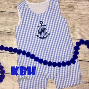 Peut inclure: Une barboteuse pour bébé en gingham bleu et blanc avec une broderie d'ancre et de monogramme. La barboteuse est sans manches et se ferme par des boutons-pression au bas. La barboteuse est sur une surface en bois avec des pompons bleus.