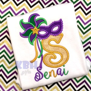 May include: A white t-shirt featuring a gold glitter letter "S" adorned with a purple and green Mardi Gras mask and a green and purple flower. The name "Senai" is embroidered in purple beneath the letter.