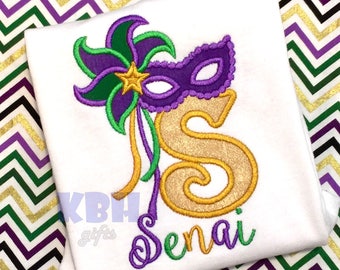 May include: A white t-shirt with a gold glitter letter "S" decorated with a purple and green Mardi Gras mask and a green and purple flower. The name "Senai" is embroidered in purple below the letter.