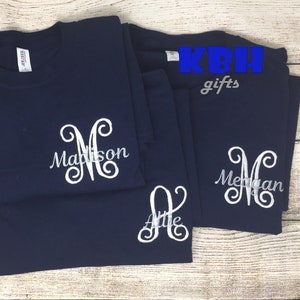 May include: Three navy blue t-shirts with white embroidered monograms. The monograms are "M" for Madison, "A" for Allie, and "M" for Megan. The shirts are folded and arranged on a wooden surface. The text "KBH gifts" is visible in the top right corner of the image.