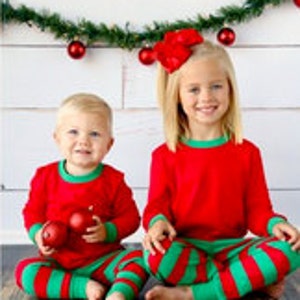 May include: Two children wearing matching red and green striped pajamas. The pajamas have a red long-sleeved shirt with green trim and green and red striped pants.