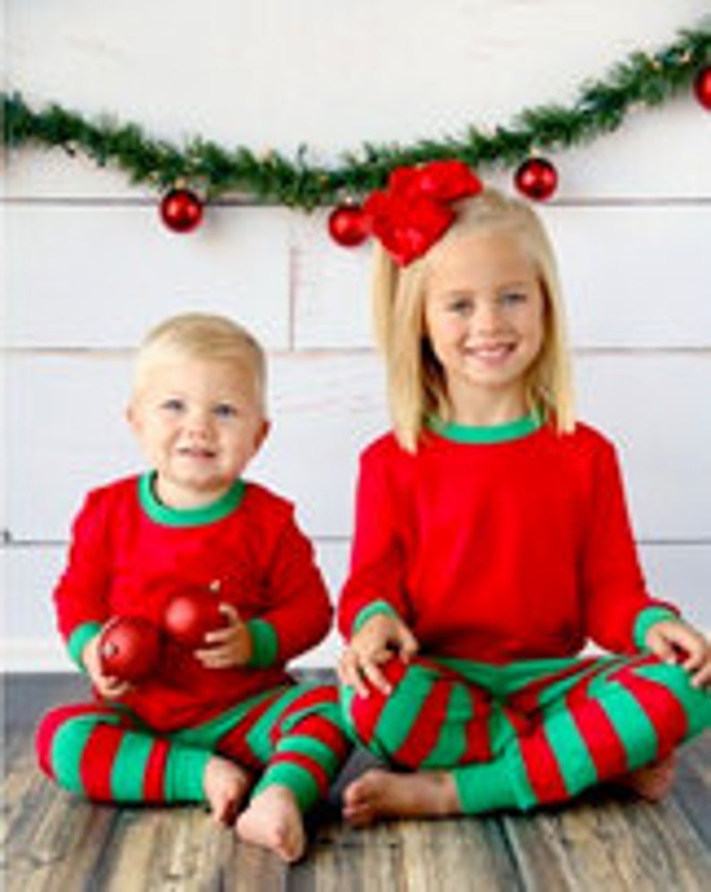 May include: Two children wearing matching red and green striped pajamas. The pajamas have a red long-sleeved shirt with green trim and green and red striped pants.