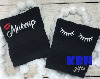May include: Two black washcloths with white embroidery. One washcloth has the words "Makeup" embroidered on it with a red lip design. The other washcloth has two white eyelashes embroidered on it. The washcloths are on a white background with white pom-poms.