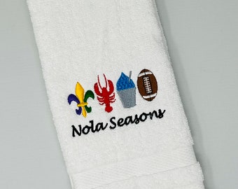 Puede incluir: Toalla de mano blanca con imágenes bordadas de un lirio, un cangrejo de río, un cono de nieve y una pelota de fútbol. El texto "Nola Seasons" está bordado debajo de las imágenes.