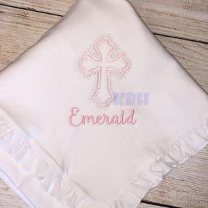 Puede incluir: Una manta de beb&eacute; blanca con una cruz bordada en rosa y el nombre "Emerald" debajo.