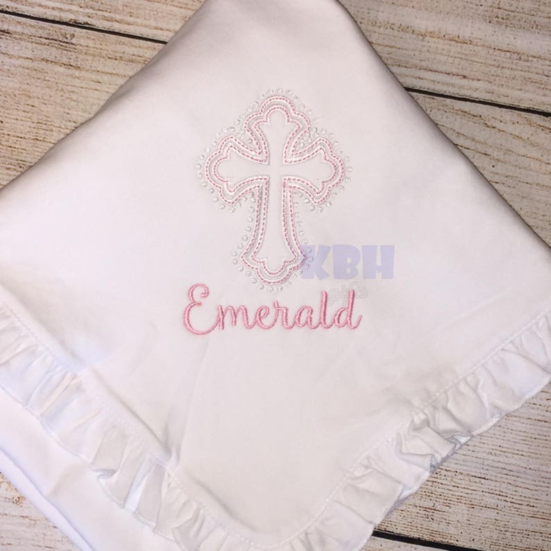 Puede incluir: Una manta de beb&eacute; blanca con una cruz bordada en rosa y el nombre "Emerald" debajo.