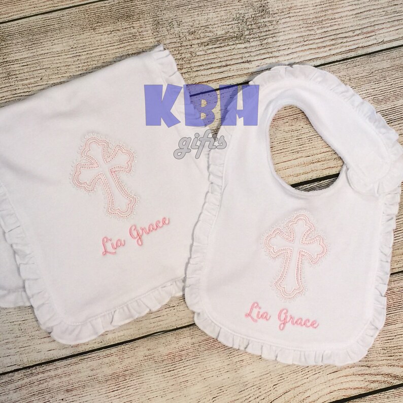 Puede incluir: Un babero y un pa&ntilde;o de eructos para beb&eacute; blancos con una cruz bordada en rosa y el nombre "Lia Grace".
