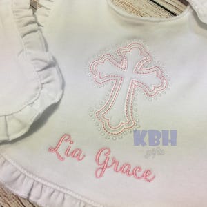 Puede incluir: Babero blanco con una cruz bordada en rosa y el nombre "Lia Grace" sobre un fondo blanco.